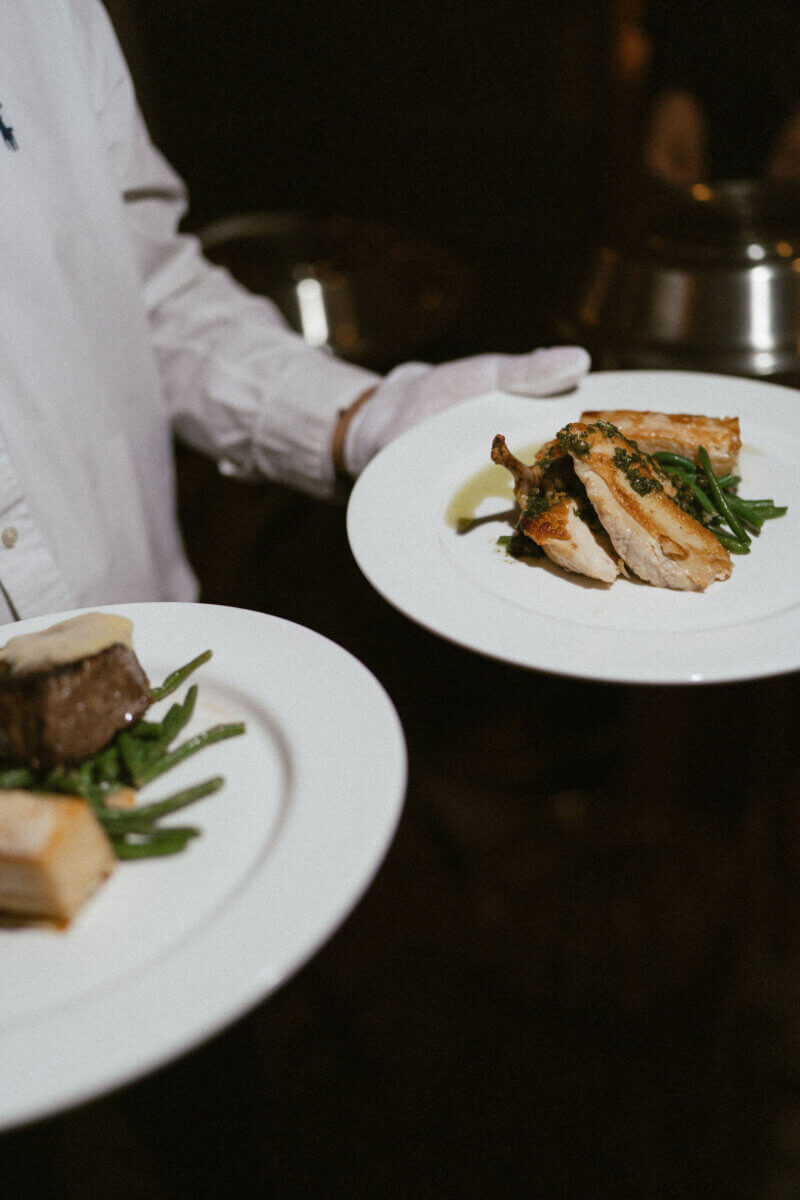 A Country Club Wedding with Show-Stopping Bridal Fashion & Peonies for Days - 21 A server in a white shirt and gloves holds two plates of food, each with green beans and a protein, one possibly chicken and the other beef—capturing the elegant dining experience typical of a classic wedding at the Belle Meade Country Club.