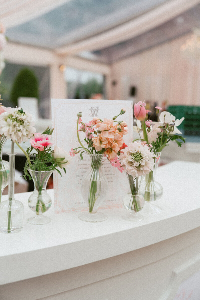 A Country Club Wedding with Show-Stopping Bridal Fashion & Peonies for Days - 13 Five small glass vases with assorted pink and white flowers are arranged on a white curved counter indoors, creating a delicate centerpiece perfect for a classic wedding. A printed menu or sign is displayed behind the vases, reminiscent of elegant gatherings at Belle Meade Country Club in Nashville.