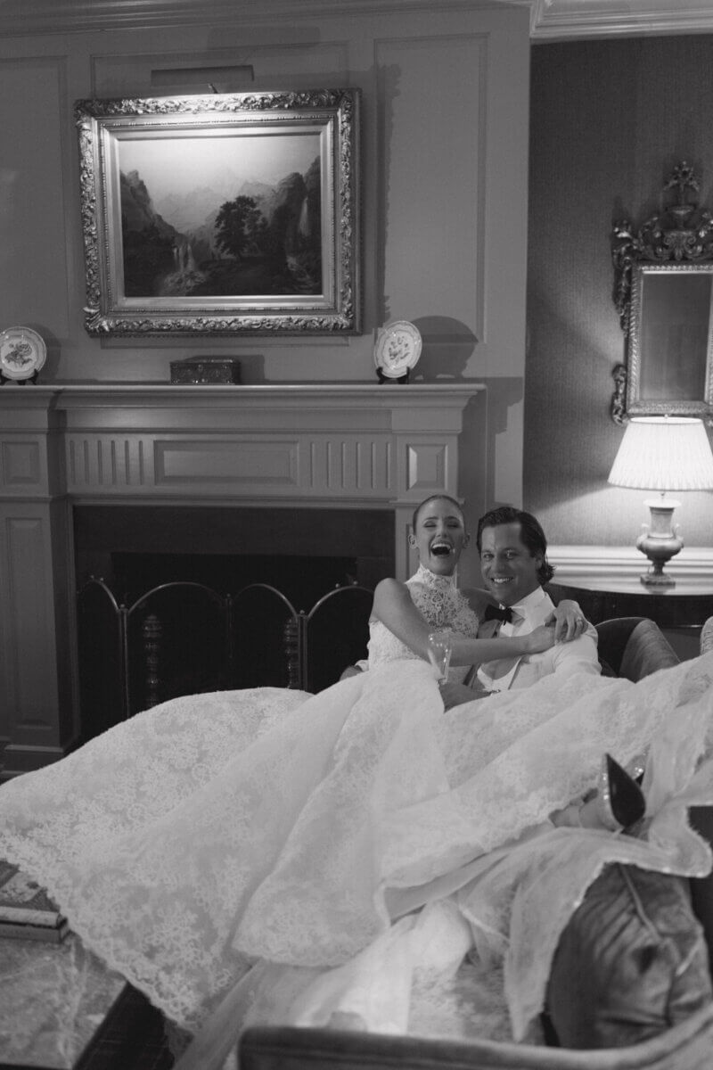 A Country Club Wedding with Show-Stopping Bridal Fashion & Peonies for Days - 7 A bride and groom in formal attire sit together on a couch, smiling and relaxed in an elegantly decorated room with classic furniture and framed art, capturing the timeless charm of a classic wedding at Belle Meade Country Club.