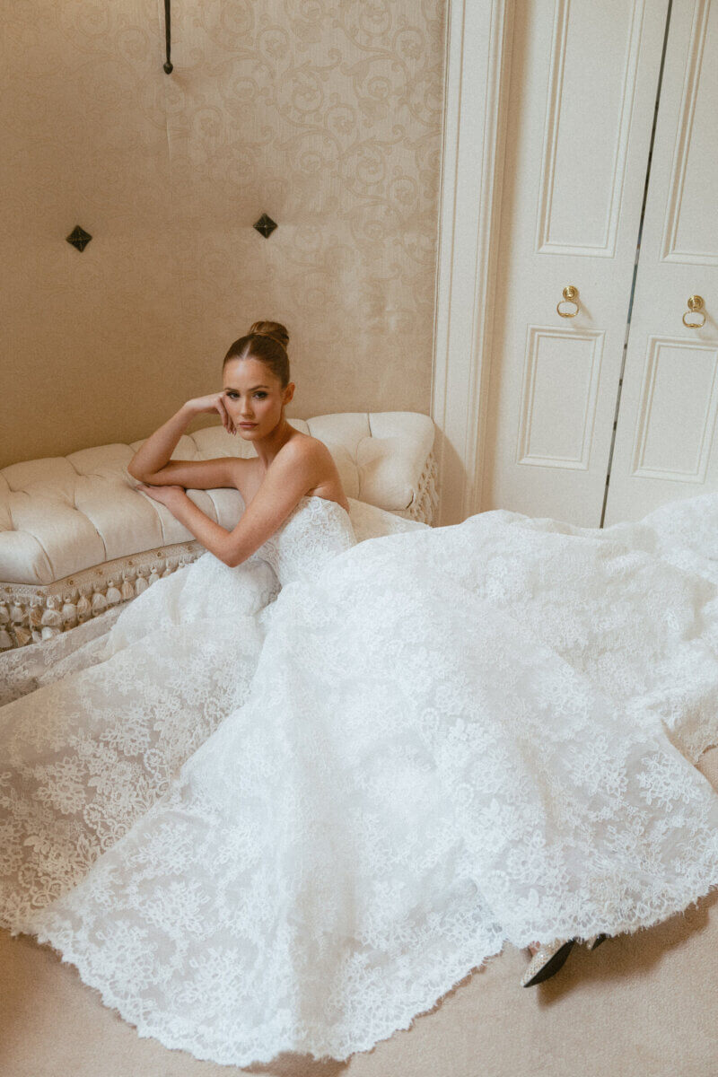 A Country Club Wedding with Show-Stopping Bridal Fashion & Peonies for Days - 5 A woman in a strapless white lace wedding dress sits on the floor, leaning against a cream-colored tufted bench in an elegant room with light walls and double doors, capturing the timeless charm of a classic wedding at Belle Meade Country Club in Nashville.