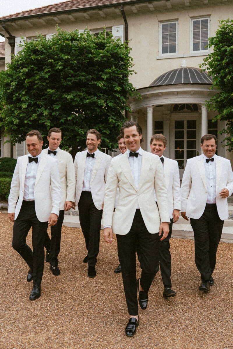 A Country Club Wedding with Show-Stopping Bridal Fashion & Peonies for Days - 8 Six men in white tuxedo jackets, black pants, and bow ties walk together outside a large house with white columns and green trees, capturing the timeless elegance of a classic wedding at Belle Meade Country Club.