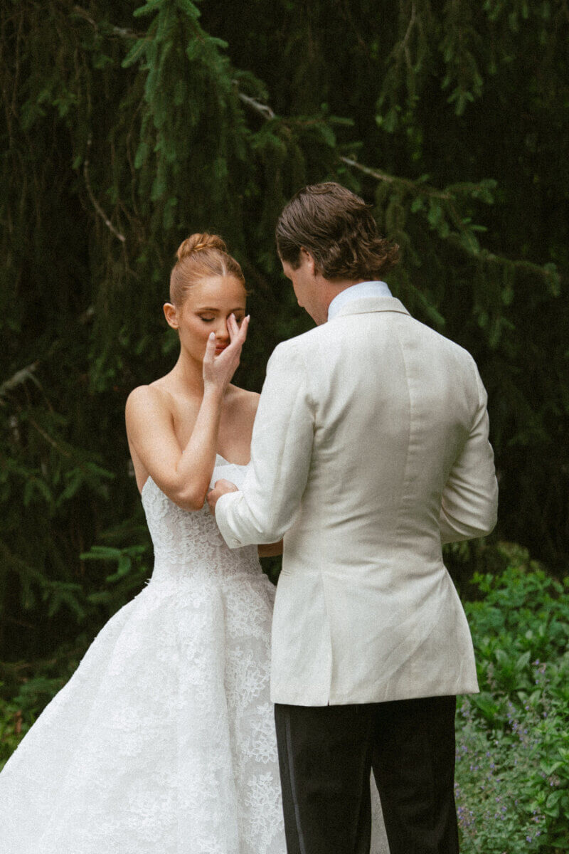 A Country Club Wedding with Show-Stopping Bridal Fashion & Peonies for Days - 14 A bride in a white lace gown stands with a groom in a white jacket in a garden at Belle Meade Country Club. She touches her face, and they hold hands, both facing each other amid the lush greenery—a classic wedding scene perfect for a Nashville wedding.