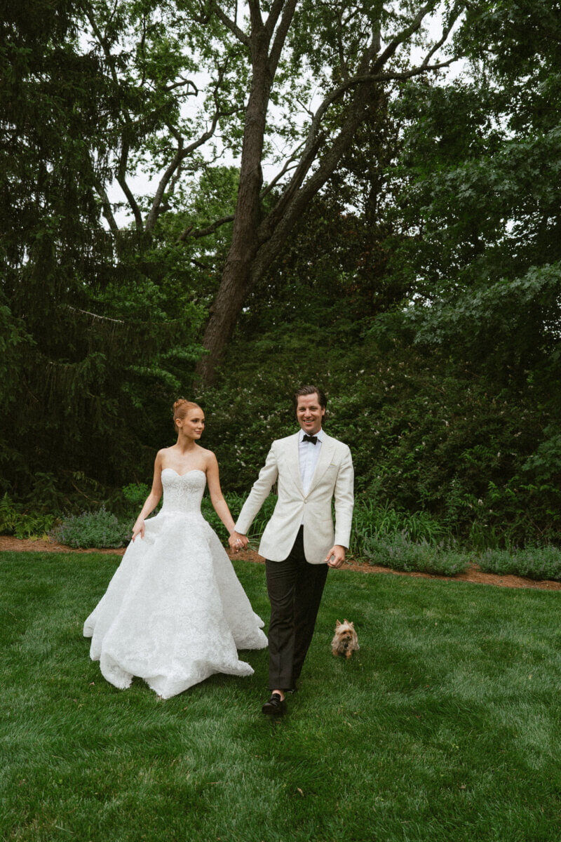 A Country Club Wedding with Show-Stopping Bridal Fashion & Peonies for Days - 15 A bride and groom walk hand in hand on a grassy lawn at Belle Meade Country Club, dressed in formal attire, with a small dog beside them and trees in the background—a perfect moment from a classic Nashville wedding.