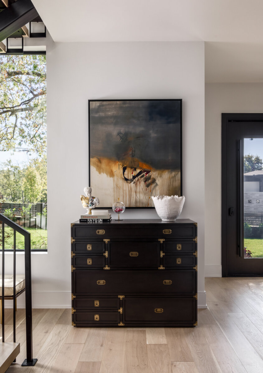 There's a Graffiti-Covered Bust in This Nashville Home, and It Works - 2 Black chest with gold accents against a white wall, topped with decor and a modern abstract painting; the space evokes a striking modern art gallery atmosphere. Large windows and a door provide ample natural light, making this Nashville home both elegant and inviting.