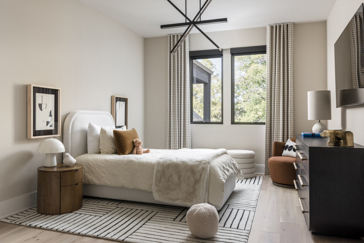 There's a Graffiti-Covered Bust in This Nashville Home, and It Works - 16 Modern bedroom with a white bed, geometric rug, round nightstand, two windows with patterned curtains, and minimalist decor. A teddy bear sits on the bed, adding a touch of warmth to this striking Nashville home that resembles a modern art gallery.