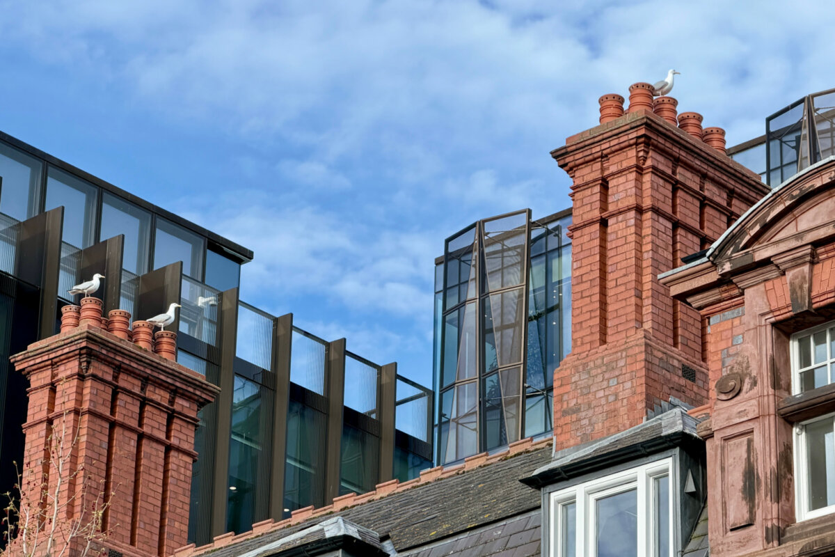 You’re About to Notice Chimney Pots Everywhere - 2 Two seagulls perch on separate red brick chimney pots of a building, with modern glass architecture and a partly cloudy blue sky in the background.