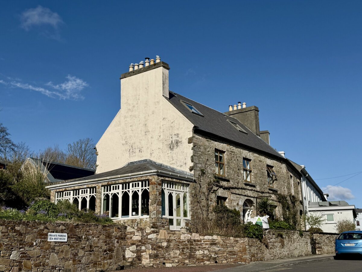 You’re About to Notice Chimney Pots Everywhere - 4 Stone house with a large white chimney adorned with classic chimney pots, slate roof, and glass conservatory under a clear blue sky. A blue car is parked on the street in front.