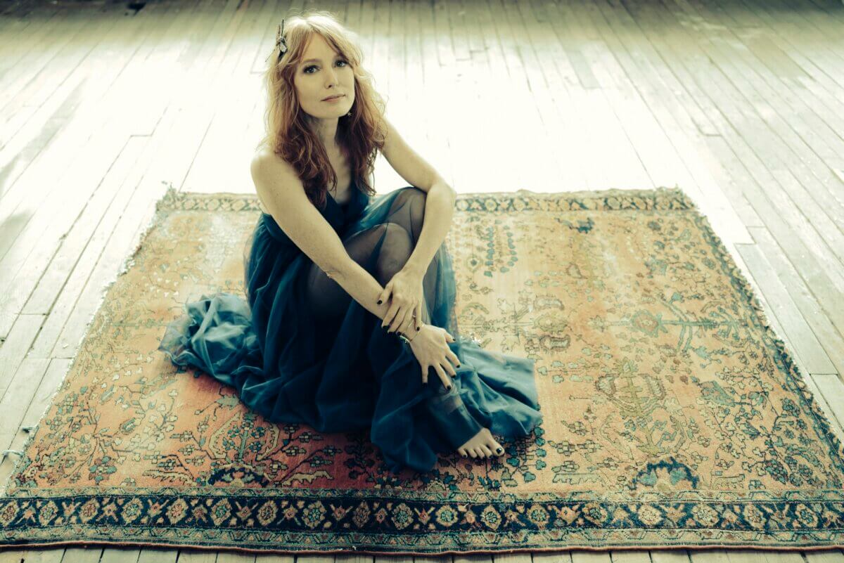 Alicia Witt Just Released Her Most Personal Album Yet - 2 Alicia Witt, in a blue dress, sits barefoot on an ornate rug atop a wooden floor, looking toward the camera in a softly lit room.