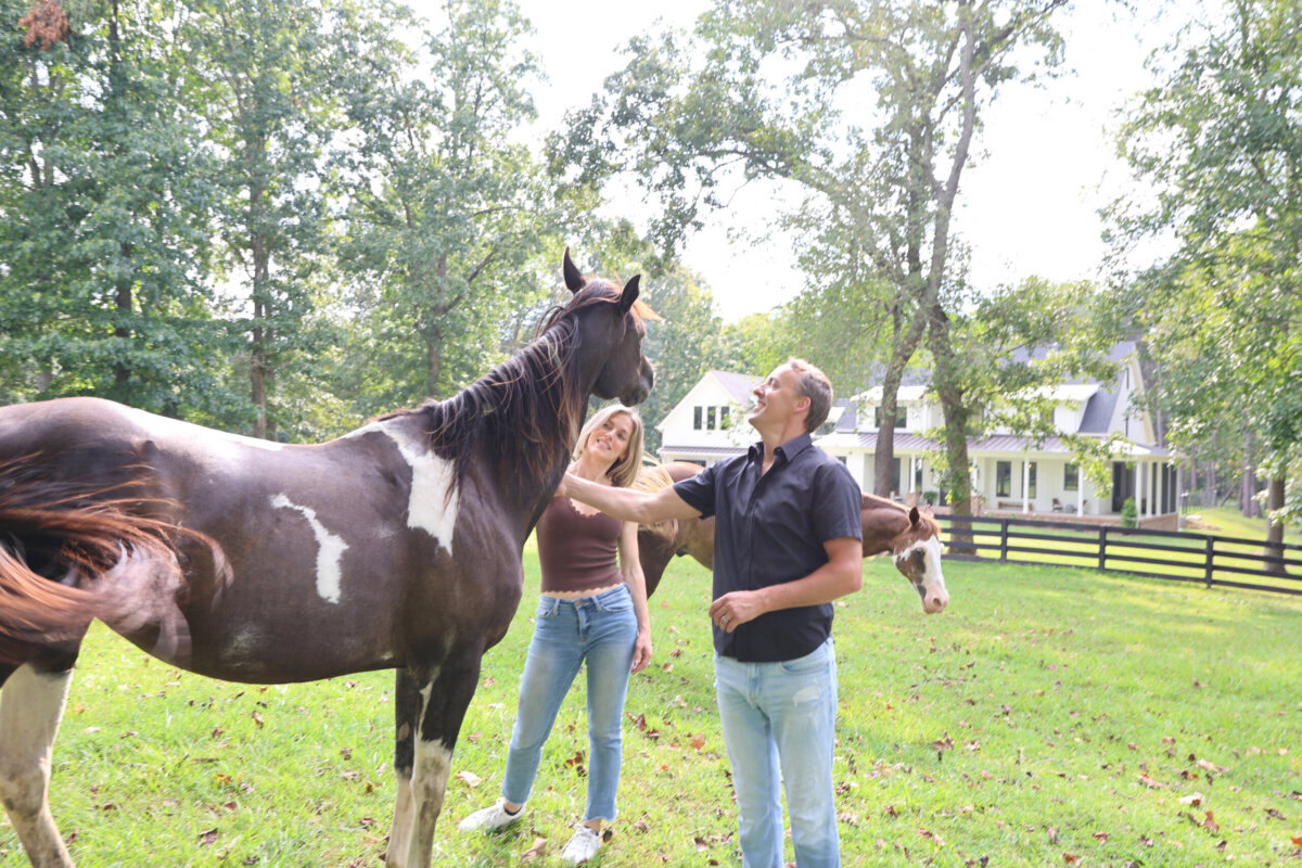 Jasmine Chambers Is Changing the Home-Building Experience - 5 Jasmine Chambers and a companion stand in a grassy yard, petting two horses, with a house and trees in the background on a sunny day.