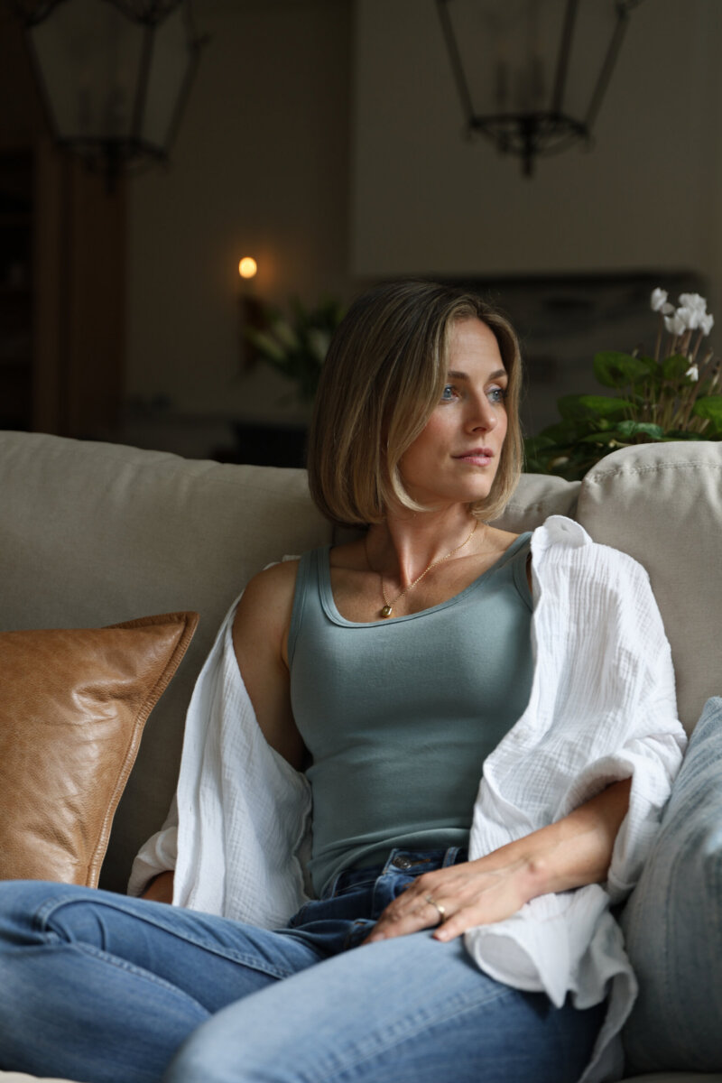 Jasmine Chambers Is Changing the Home-Building Experience - 4 Jasmine Chambers, a woman with short blonde hair, sits on a beige couch wearing a light blue tank top, white open shirt, and jeans, looking thoughtfully to the side.