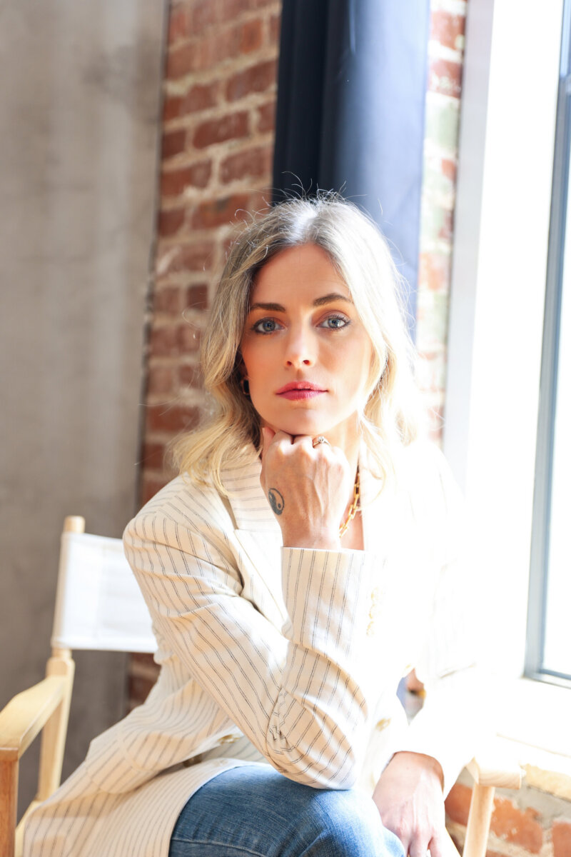 Jasmine Chambers Is Changing the Home-Building Experience - 2 Jasmine Chambers, a woman with blonde hair, wears a light pinstripe blazer and jeans as she sits on a wooden chair by a window, resting her chin on her hand and looking at the camera.
