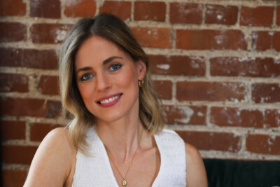 Jasmine Chambers, a woman with shoulder-length blonde hair, wearing a white sleeveless top and gold jewelry, smiles in front of a brick wall.