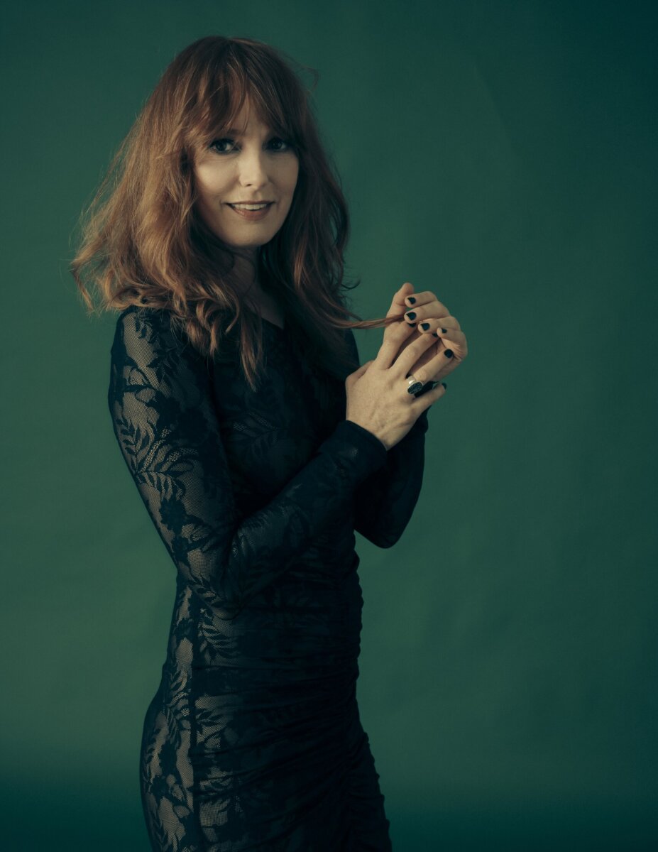 Alicia Witt Just Released Her Most Personal Album Yet - 6 Alicia Witt wears a fitted black lace dress as she stands against a dark green background, looking at the camera and holding a strand of her hair.