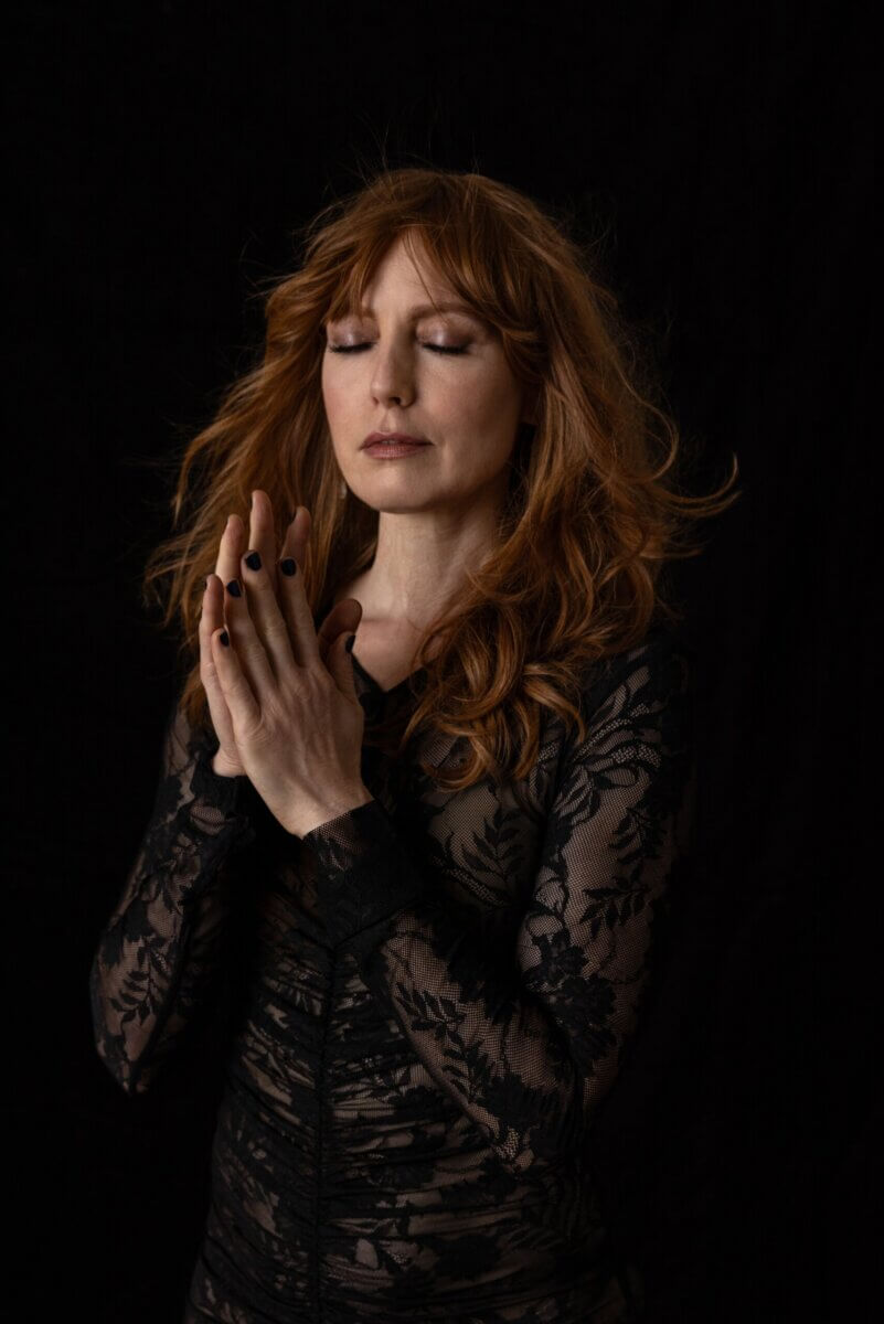 Alicia Witt Just Released Her Most Personal Album Yet - 5 Alicia Witt, a woman with long red hair and closed eyes, stands against a black background, wearing a sheer black lace top, with her hands pressed together near her face.