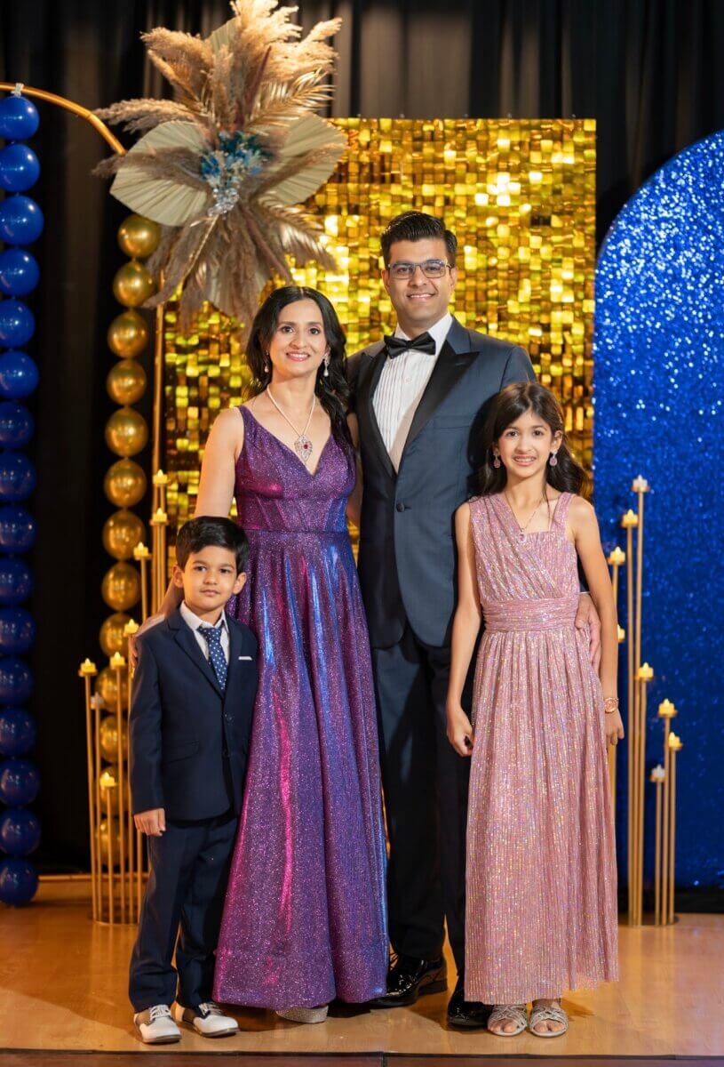 This Nashville Doctor Helps Families Disconnect to Reconnect - 5 A family of four, including Nidhi Gupta, dressed in formal attire poses in front of a backdrop decorated with gold and blue balloons and glittery panels.