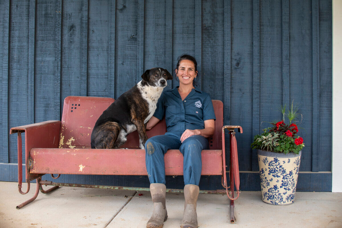 Meet the Lawyer-Turned-Farmer Behind Tennessee’s 5 Porch Farm - 3 A woman in work clothes and boots sits on a worn metal bench with her dog beside her, in front of a blue wooden wall with a flower pot on the right—an authentic moment that perfectly captures the everyday grit and warmth of Justine Moreau.