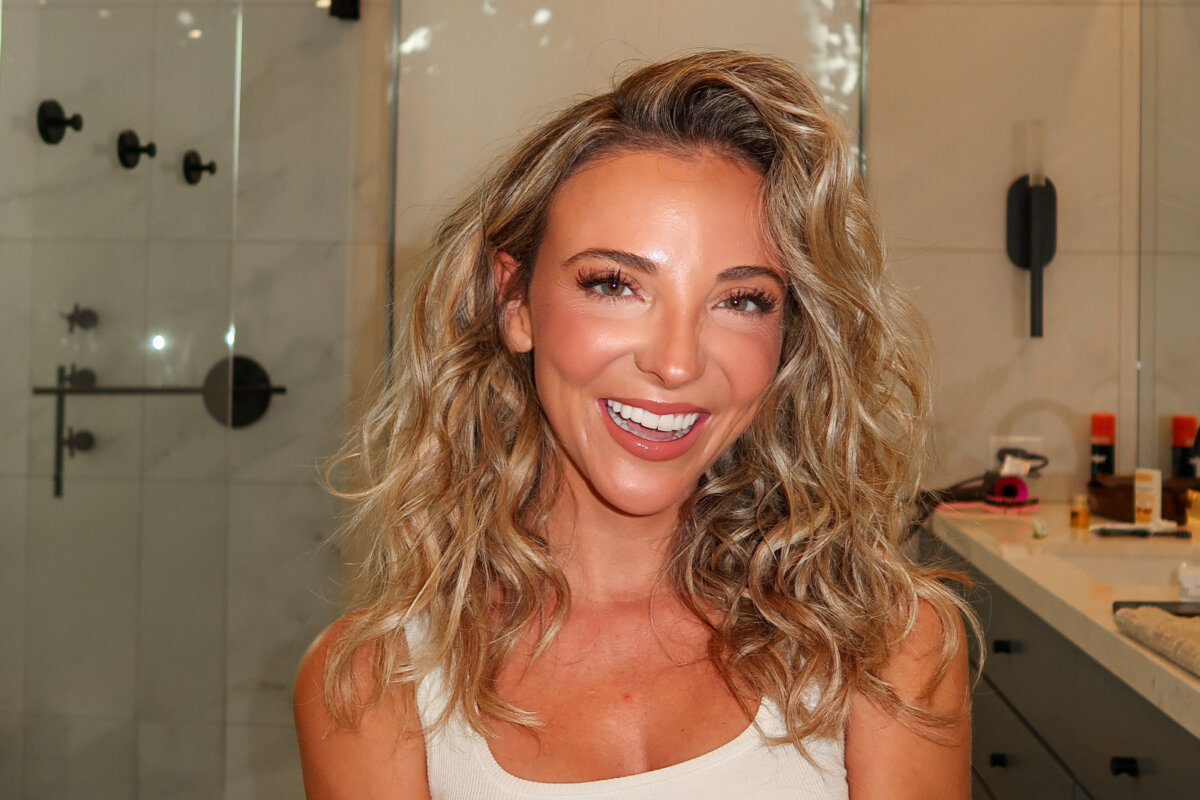 Dani Austin Is Changing the Conversation Around Hair Loss - 2 Woman with wavy blonde hair smiling in a bathroom, wearing a sleeveless top. Shower and various toiletries, including Divi hair products from Dani Austin’s collection, are visible in the background.