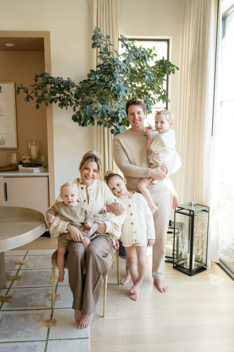 Dani Austin Is Changing the Conversation Around Hair Loss - 5 A family of five, dressed in light-colored clothing, poses together in a bright living room with large windows and a potted plant in the background, their smiles shining as they show off their healthy locks styled with Divi hair products inspired by Dani Austin.