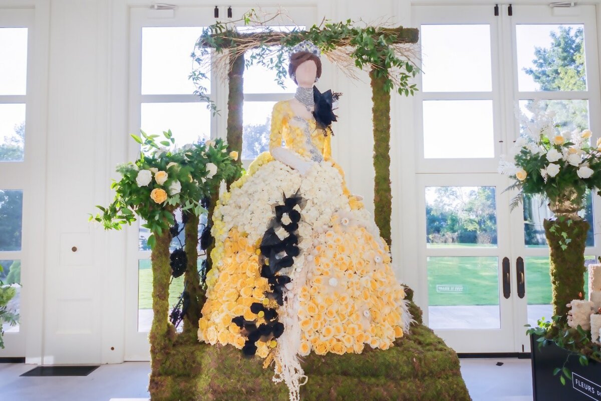 Fleurs de Villes: Cheekwood’s Latest Exhibit is a Must-See - 7 A faceless mannequin in a ballgown made of yellow and white flowers with black accents, displayed on a moss-covered platform and surrounded by floral arrangements indoors—evoking the enchanting displays often found at Cheekwood.