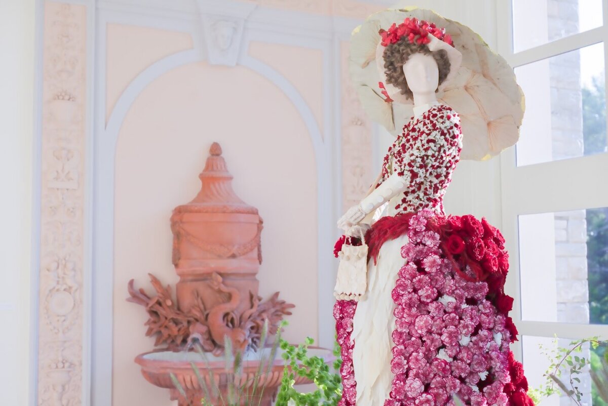 Fleurs de Villes: Cheekwood’s Latest Exhibit is a Must-See - 2 Mannequin dressed in a gown and hat decorated with red and pink flowers stands indoors near a terracotta urn and bright windows, evoking the elegant displays often found at Cheekwood.