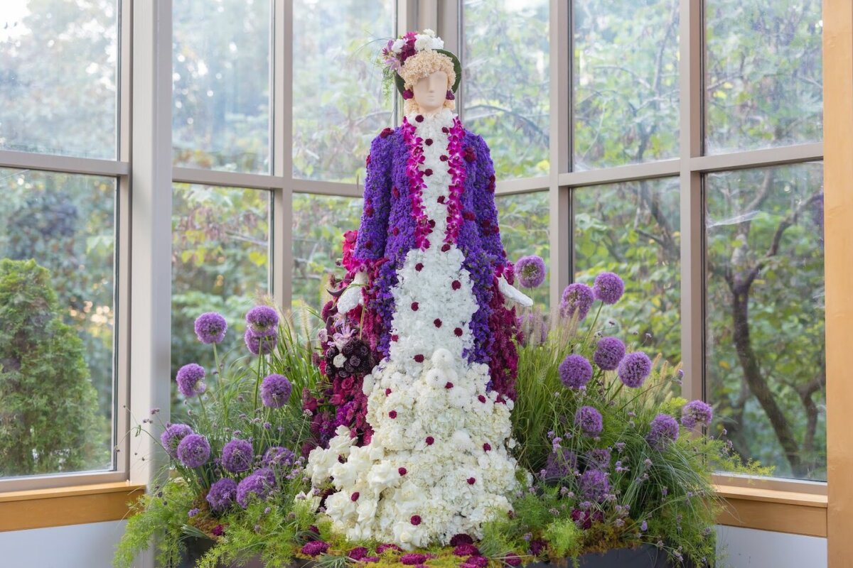 Fleurs de Villes: Cheekwood’s Latest Exhibit is a Must-See - 8 A mannequin in a garden room at Cheekwood is dressed in a gown made of purple and white flowers, surrounded by tall purple blossoms and lush greenery.