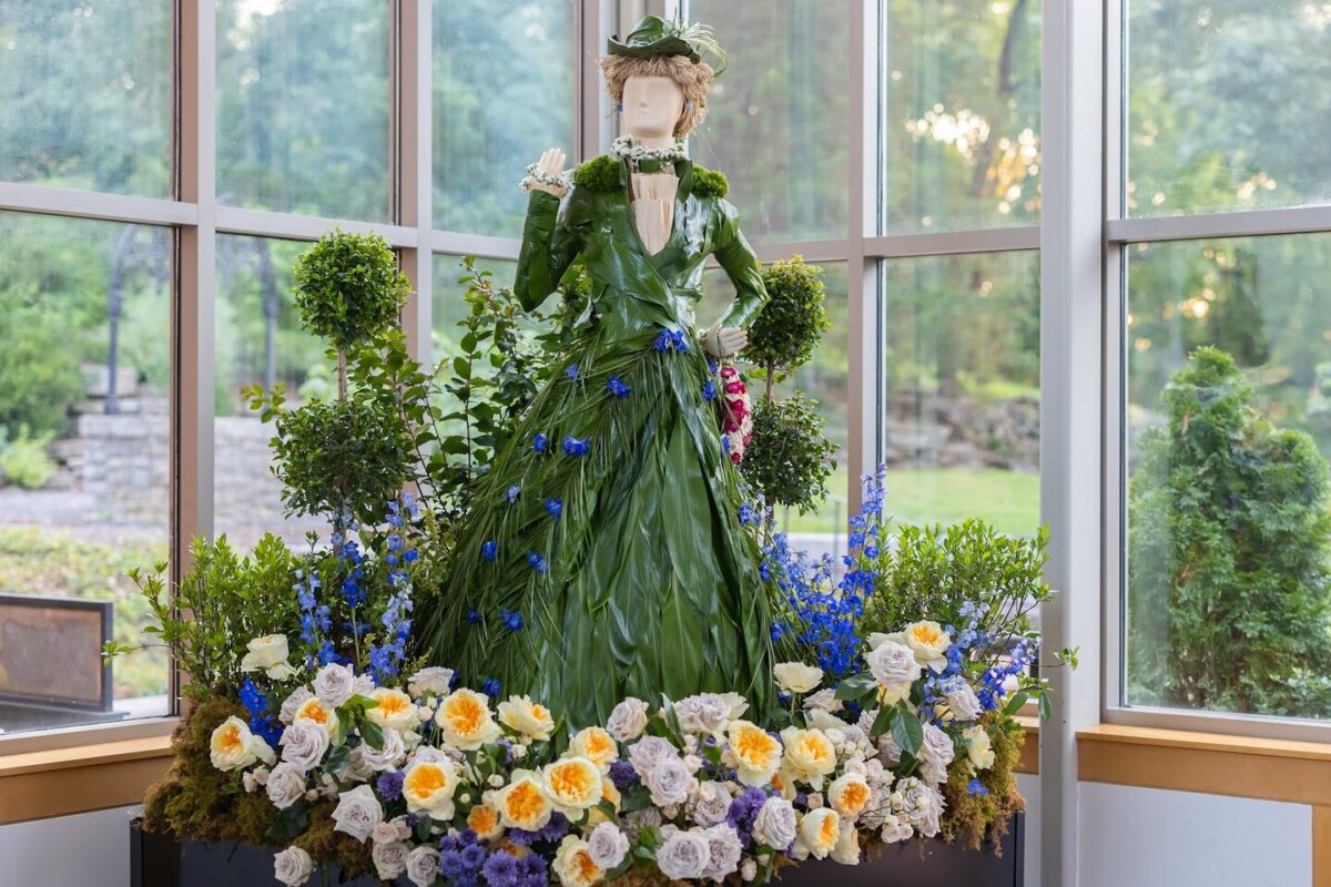 Fleurs de Villes: Cheekwood’s Latest Exhibit is a Must-See - 4 A mannequin dressed in a gown made of green leaves stands among assorted flowers inside a room with large windows overlooking a garden at Cheekwood.