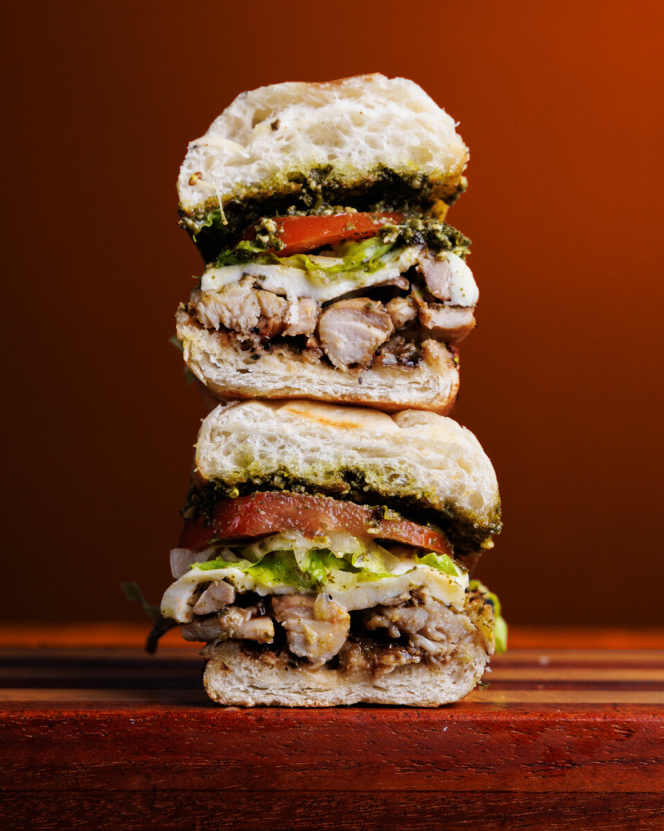 12 New Nashville Restaurants to Try This Month - 17 A stacked sandwich cut in half shows chicken, lettuce, tomato, melted cheese, and green pesto sauce between thick bread slices against an orange background—a mouthwatering preview of what to expect at new Nashville restaurants this August.