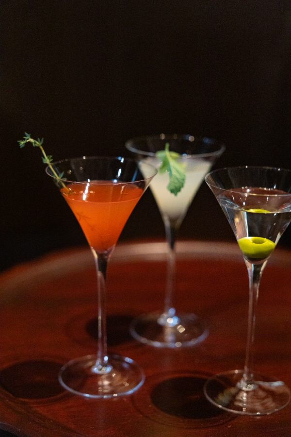 12 New Nashville Restaurants to Try This Month - 13 Three cocktails in martini glasses, each garnished differently—one with a herb sprig, one with a leafy garnish, and one with a green olive—are displayed on a wooden surface, evoking the creative spirit found in new restaurants opening across Nashville this August.