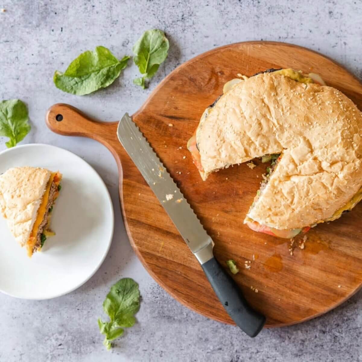 12 New Nashville Restaurants to Try This Month - 4 A round sandwich with a slice cut out sits on a wooden board next to a serrated knife; the slice is on a white plate. Green leaves are scattered on the gray surface, evoking the fresh and inviting presentations often found at new Nashville restaurants.