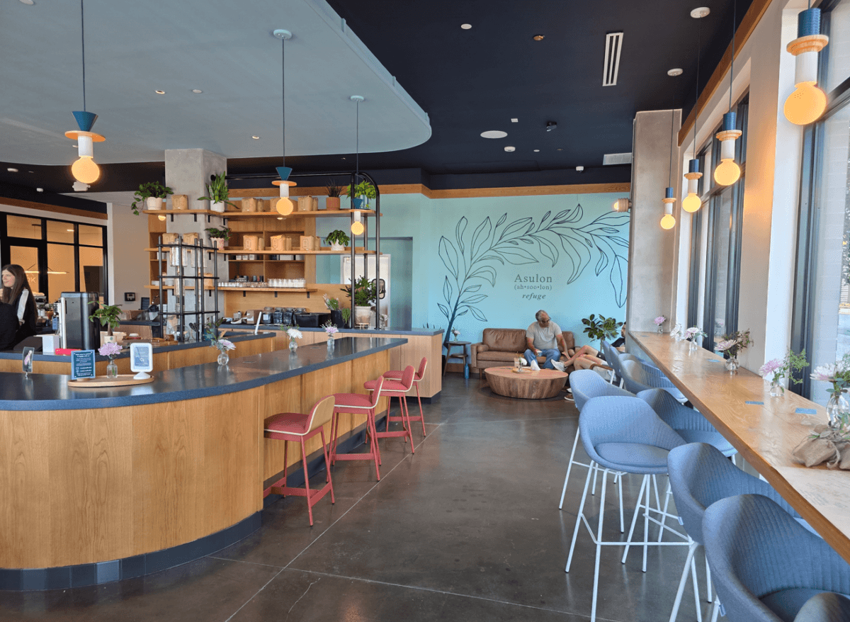 12 New Nashville Restaurants to Try This Month - 2 Modern coffee shop interior with a curved counter, bar seating, shelves with plants, and people sitting on couches near a mint green wall. Natural light streams through large windows, creating an inviting atmosphere reminiscent of the stylish designs seen in new Nashville restaurants.