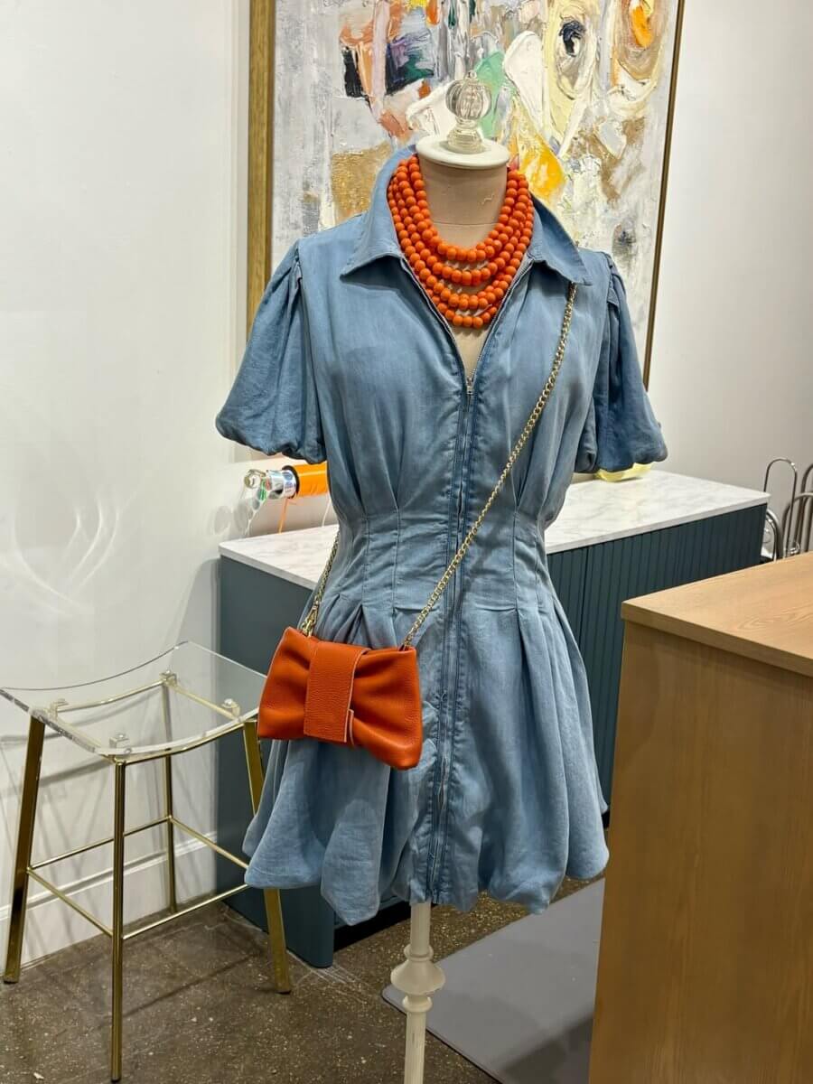 Displayed in a retail setting reminiscent of the vibrant Nashville shopping scene, this mannequin is dressed in a blue denim romper, complemented by an orange beaded necklace and a small orange purse with a chain strap—a chic ensemble inspired by the stylish offerings found in top Nashville stores.
