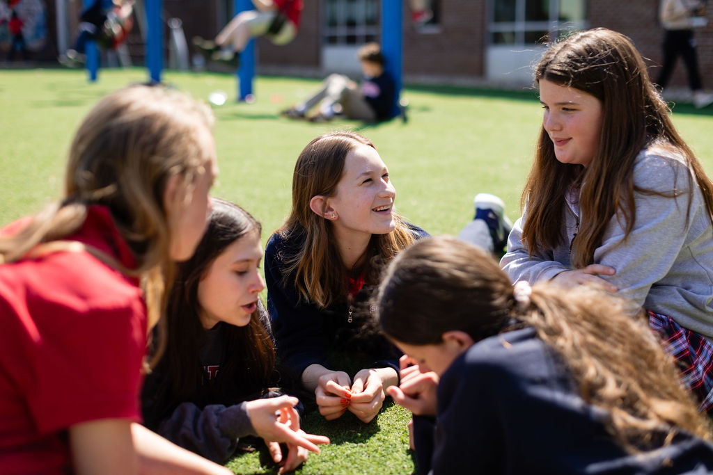Nashville Private School Open House Dates - 12 Five girls sit in a circle on artificial grass outdoors at one of the local private schools, talking and smiling, with playground equipment and other children visible in the background.