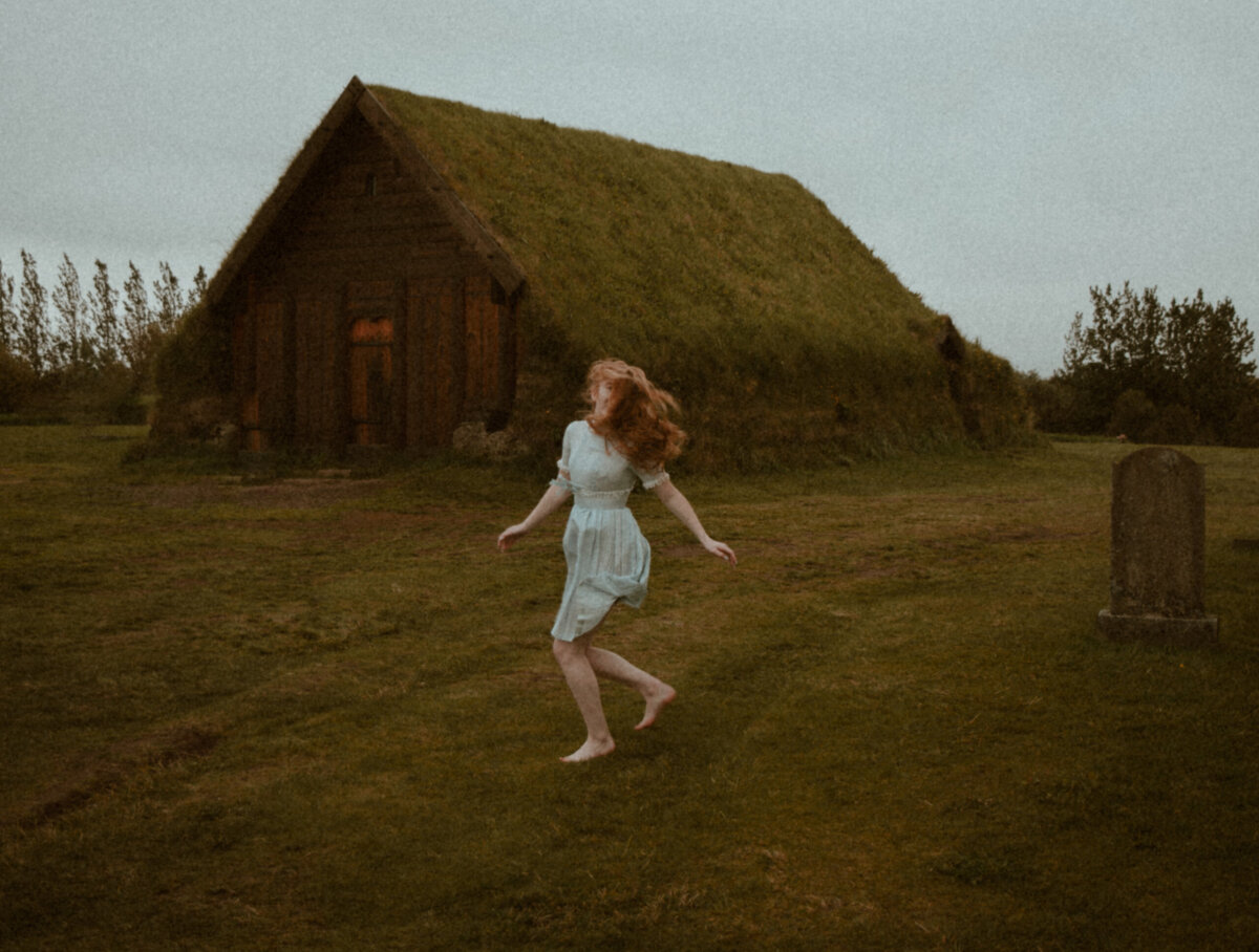 A barefoot woman in a light dress runs across grass near a turf-roofed wooden house and a gravestone, under a cloudy sky.