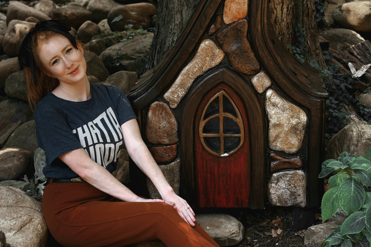 A woman sits on rocks next to a whimsical miniature stone and wood house, built against a tree trunk in a wooded area.