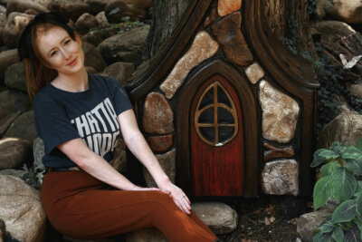 A woman sits on rocks beside a small, decorative wooden house structure with a red door and stone-like exterior, surrounded by greenery—capturing the whimsical charm often found in Chattanooga tourism spots.
