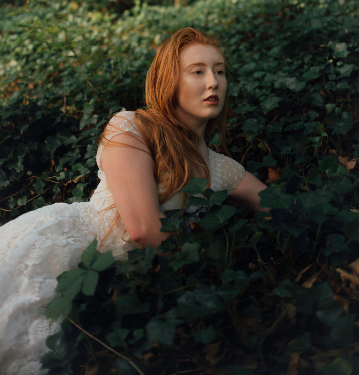 A woman with long red hair in a white dress lies among dense green ivy outdoors in Chattanooga, her gaze fixed thoughtfully into the distance.