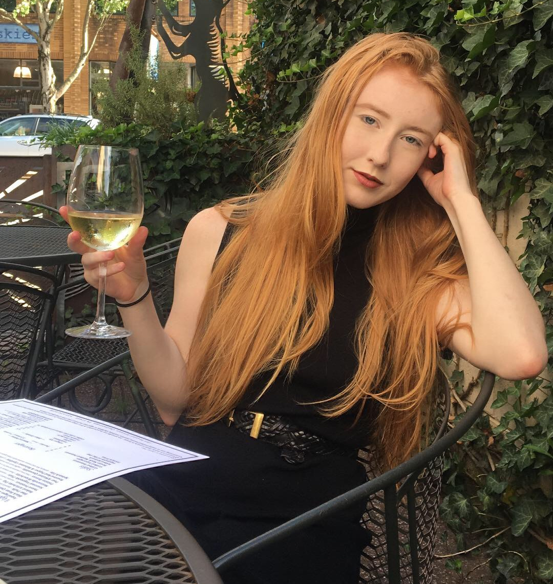 A woman with long red hair in a black dress sits at an outdoor table in Chattanooga, holding a glass of white wine, with a menu on the table and ivy in the background.