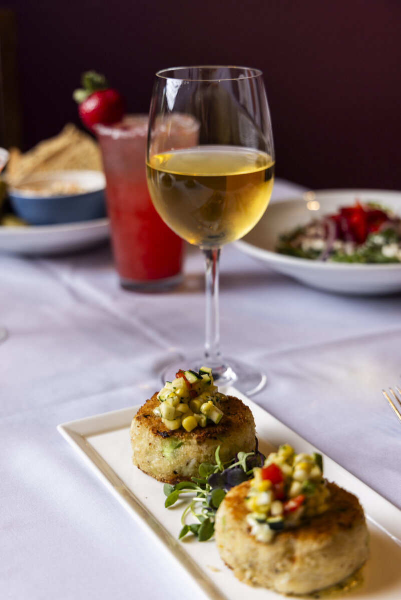 5 Destinations Where Southern Charm Thrives - 7 A glass of white wine with two crab cakes topped with corn salsa sits elegantly on a rectangular plate, set on a white tablecloth with food and drinks in the background.