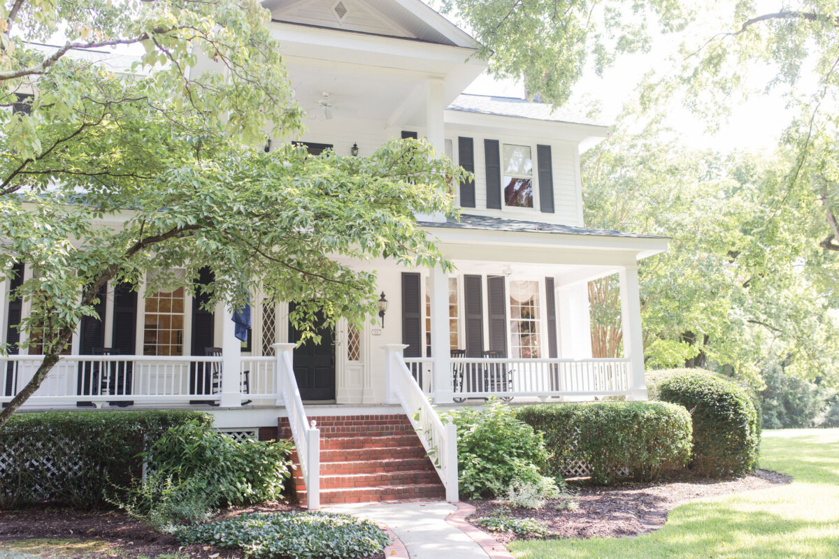 5 Destinations Where Southern Charm Thrives - 2 Two-story white house with a wraparound porch, black shutters, and brick steps, surrounded by green trees and bushes on a sunny day.