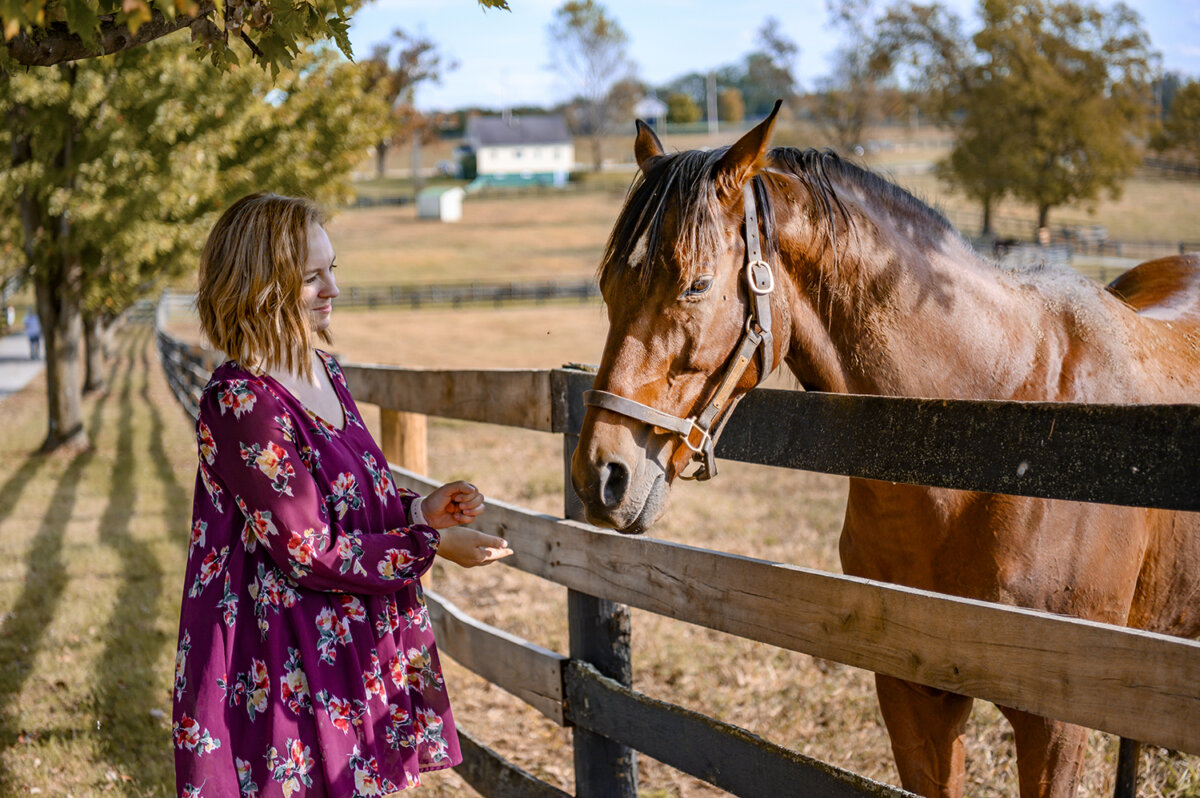 5 Destinations Where Southern Charm Thrives - 8 A woman in a floral dress stands by a wooden fence, reaching out to a brown horse in a rural field.