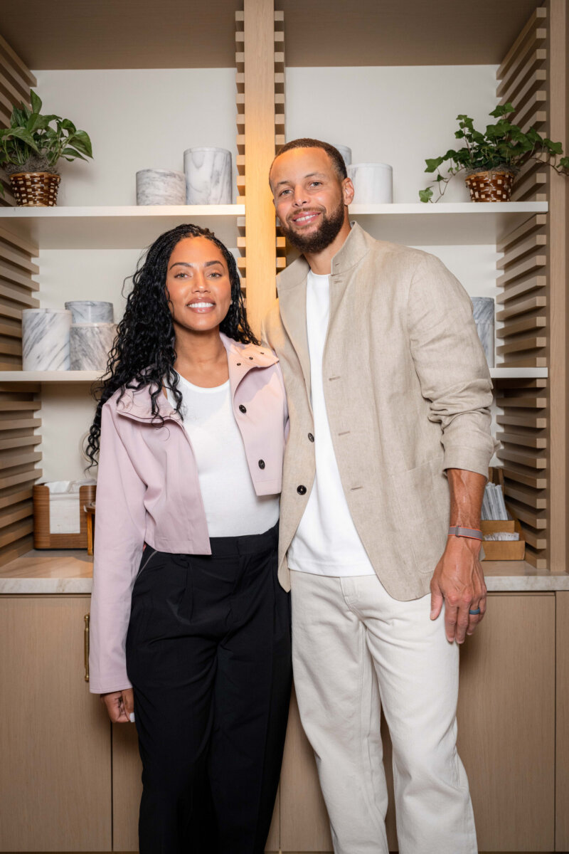 Two people stand side by side in front of wooden shelves with potted plants and marble decor, smiling at the camera.