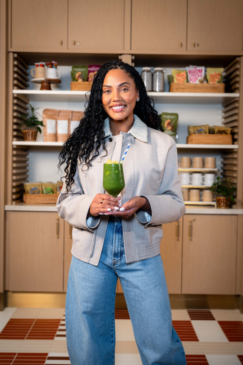 Woman standing in a kitchen holding a green smoothie with a striped straw, smiling at the camera. Shelves with food items are visible in the background.