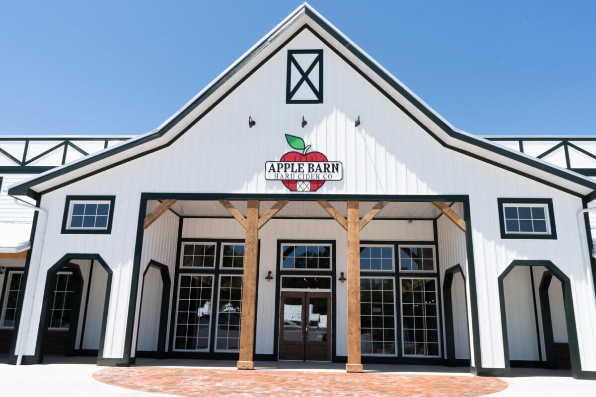 Exterior view of a white barn-style building with wood beams, large windows, and a sign that reads “Apple Barn Hard Cider Co.” against a clear blue sky—a favorite stop for those looking to raise a glass and savor authentic Southern spirits.