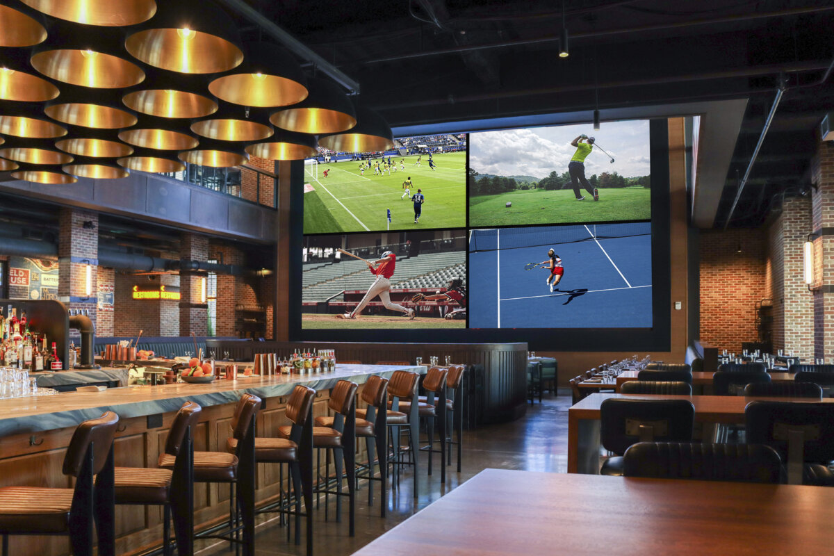 A modern sports bar with a large screen displaying football, golf, baseball, and tennis, surrounded by wooden tables, chairs, and a fully stocked bar offering an impressive selection of southern spirits.