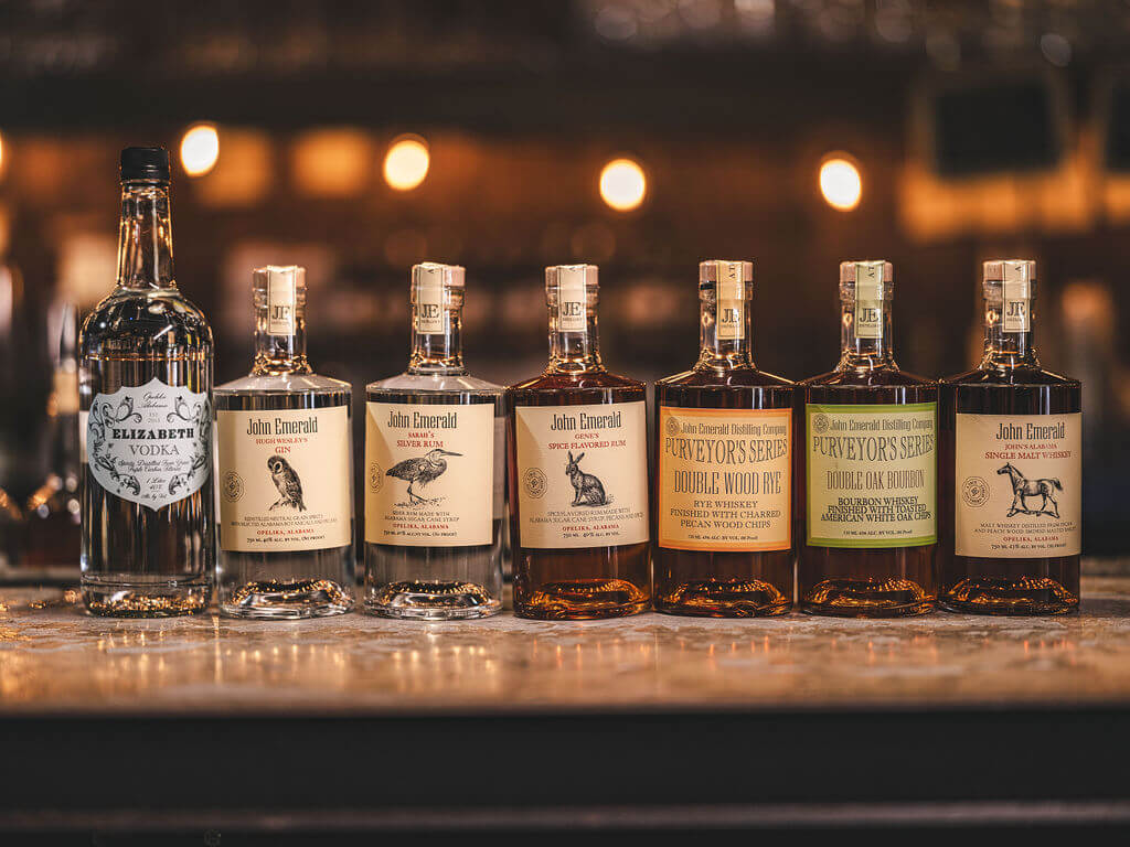Seven bottles of liquor, including vodka and various whiskeys, are lined up on a bar counter with a blurred background—perfect for those ready to Raise A Glass to their Favorite Stops featuring the finest Southern Spirits.