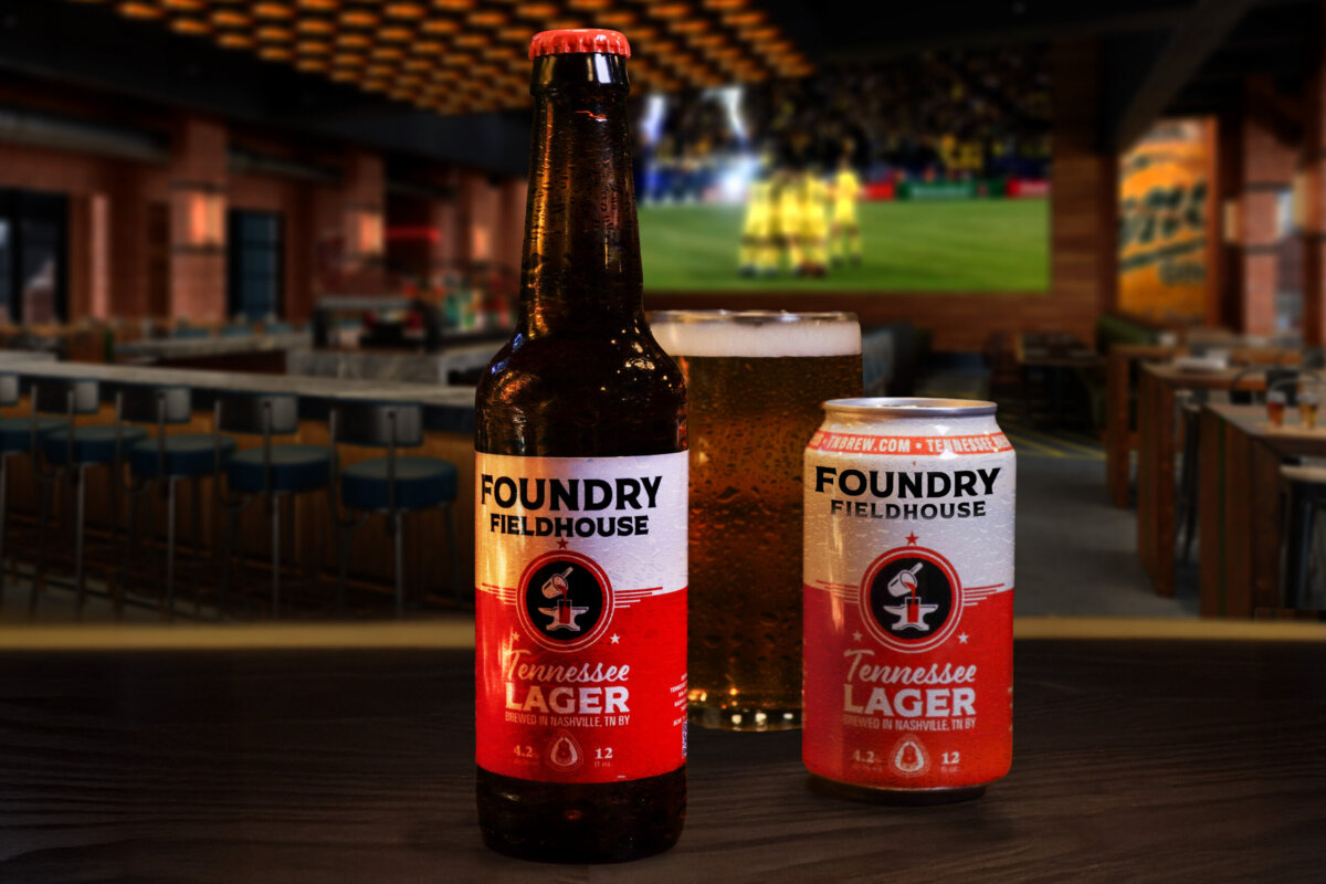 A Foundry Fieldhouse Tennessee Lager bottle, can, and a beer glass sit on a bar counter with a blurred sports bar and a large TV screen in the background, capturing the lively essence of southern spirits.