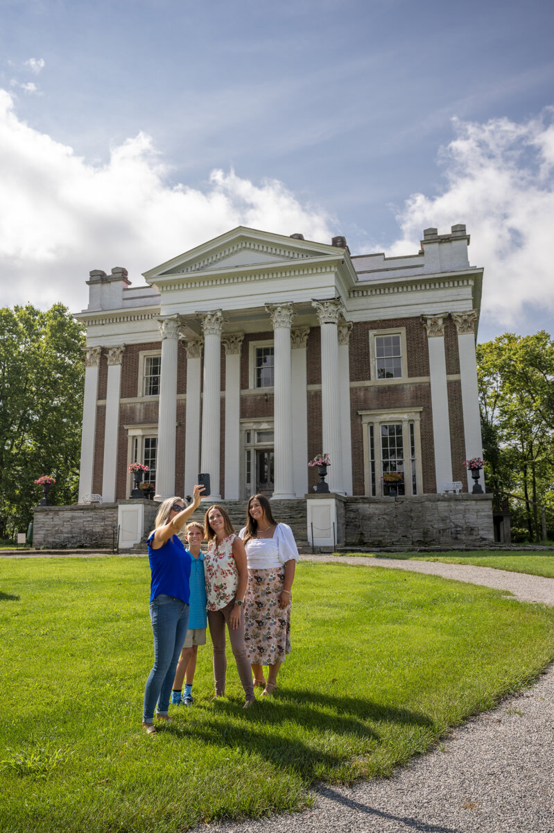 5 Destinations Where Southern Charm Thrives - 9 Four people stand on a lawn taking a selfie in front of a large historic mansion with tall white columns on a sunny day, capturing the essence of Southern charm at one of the most iconic travel destinations in the Southern USA.