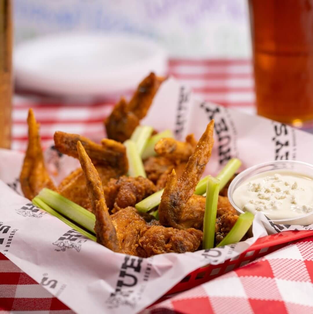 A basket of fried chicken wings with celery sticks and a cup of ranch dip sits on a red and white checkered tablecloth.