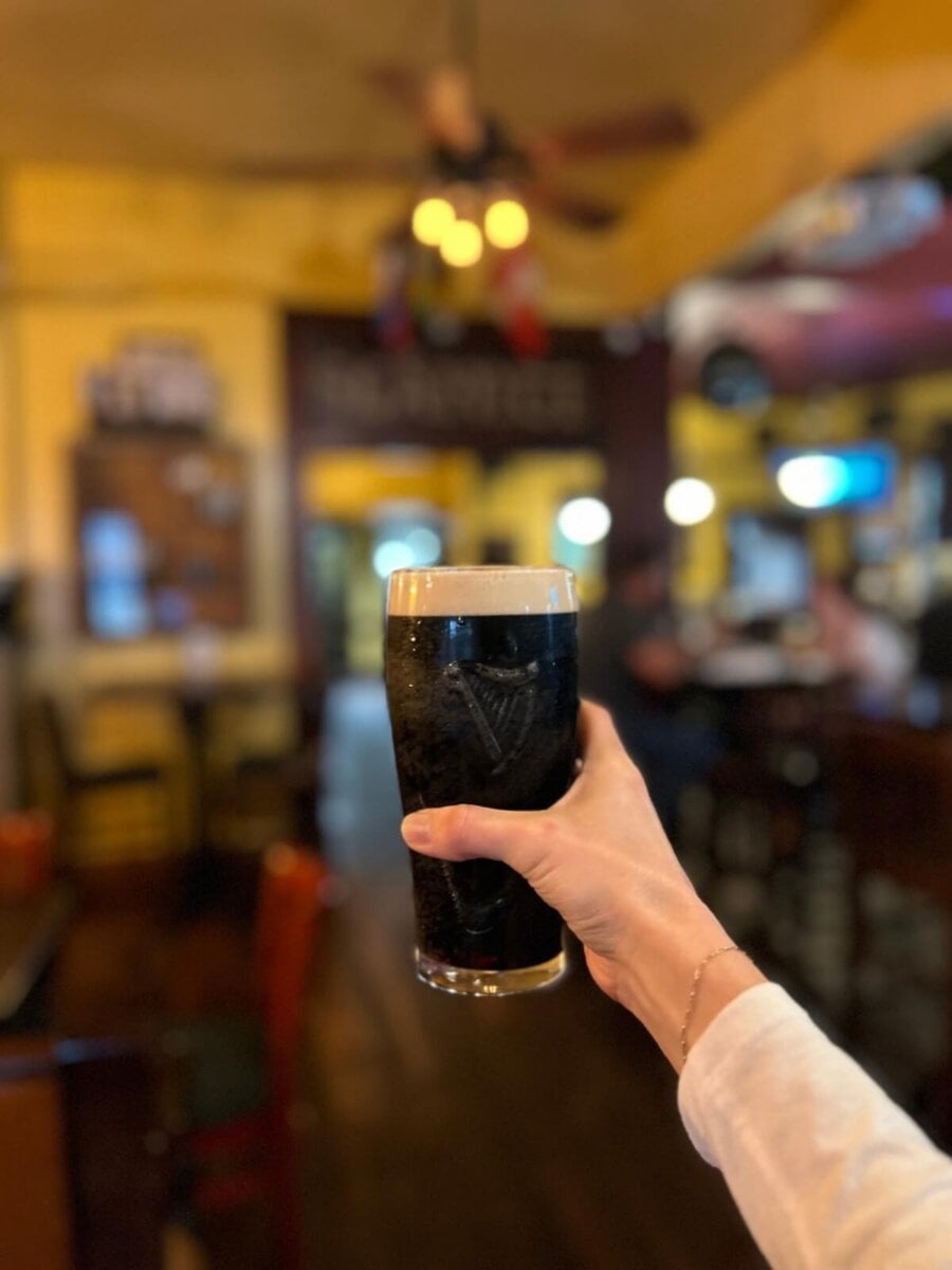 A hand holding a pint of dark beer in a glass, with a blurred view of a pub interior in the background.