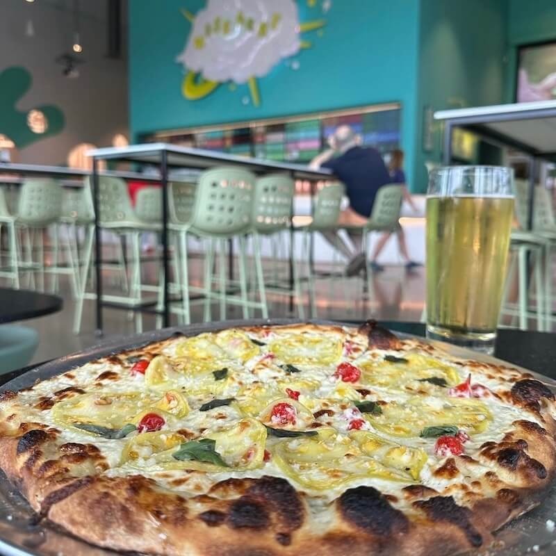 A pizza with yellow tomatoes, cherry tomatoes, and basil sits on a table next to a glass of beer in a modern, brightly lit restaurant.